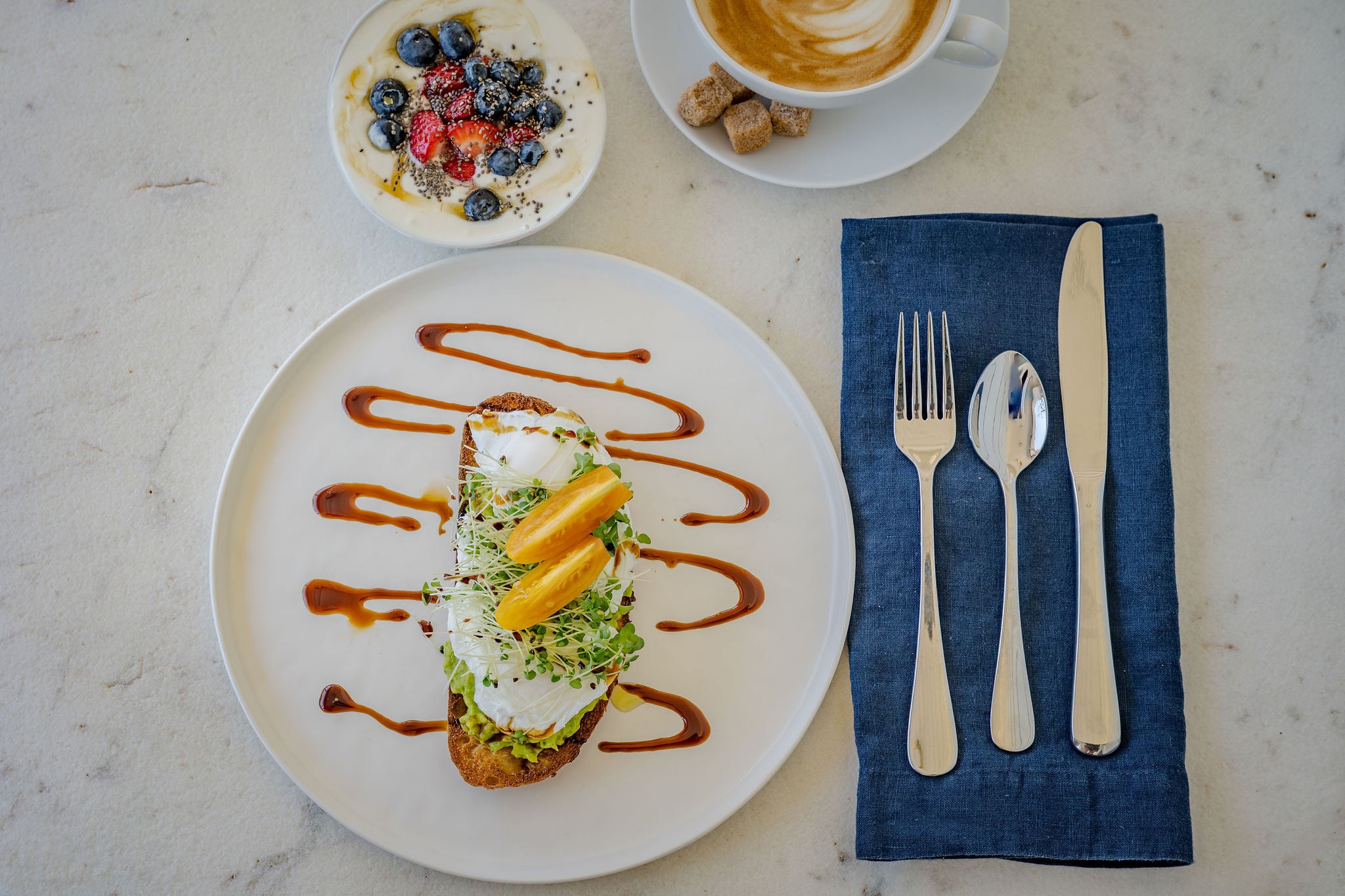 Avocado toast with a poached egg, a berry yogurt bowl and a latte laid out on marble